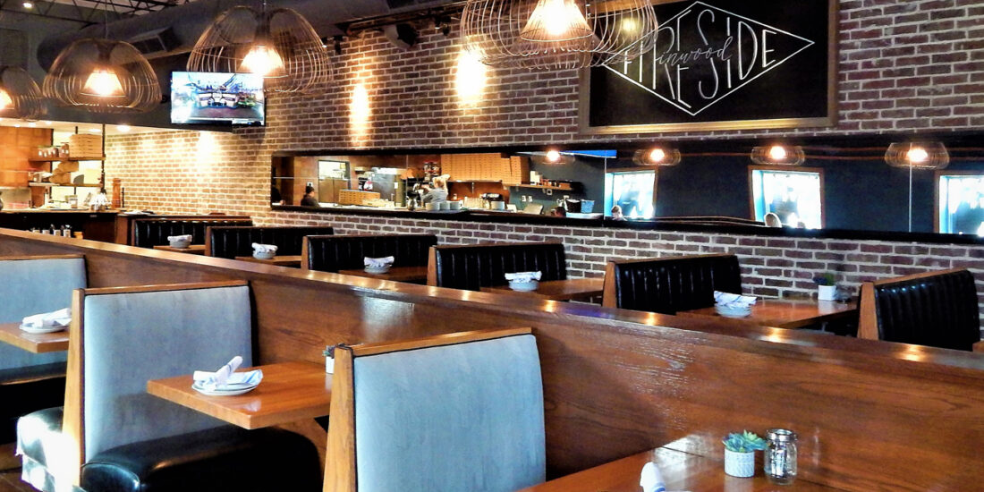 Fireside Pies restaurant interior with a cozy, rustic ambiance, featuring wooden tables, blue suede and leather booth seating, dim lighting, and an open kitchen showcasing a brick pizza oven.
