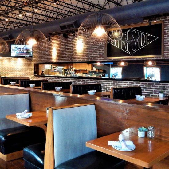 Fireside Pies restaurant interior with a cozy, rustic ambiance, featuring wooden tables, blue suede and leather booth seating, dim lighting, and an open kitchen showcasing a brick pizza oven.